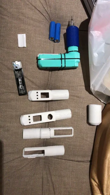 Carcaças brancas impressas, corpo verde-água montado com empunhadura azul, duas baterias e uma placa eletrônica sobre um estofado